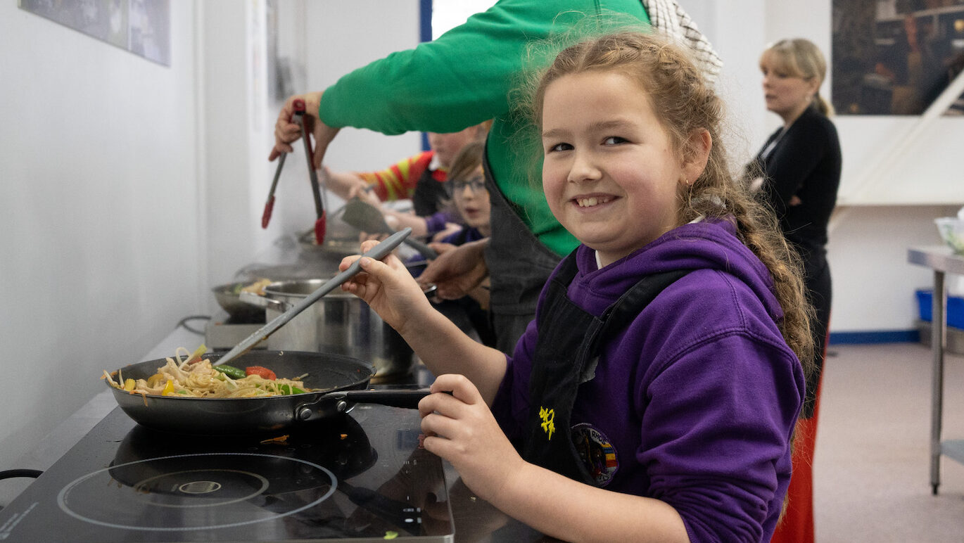 child looks at camera, cooking in a frying pan on a hob